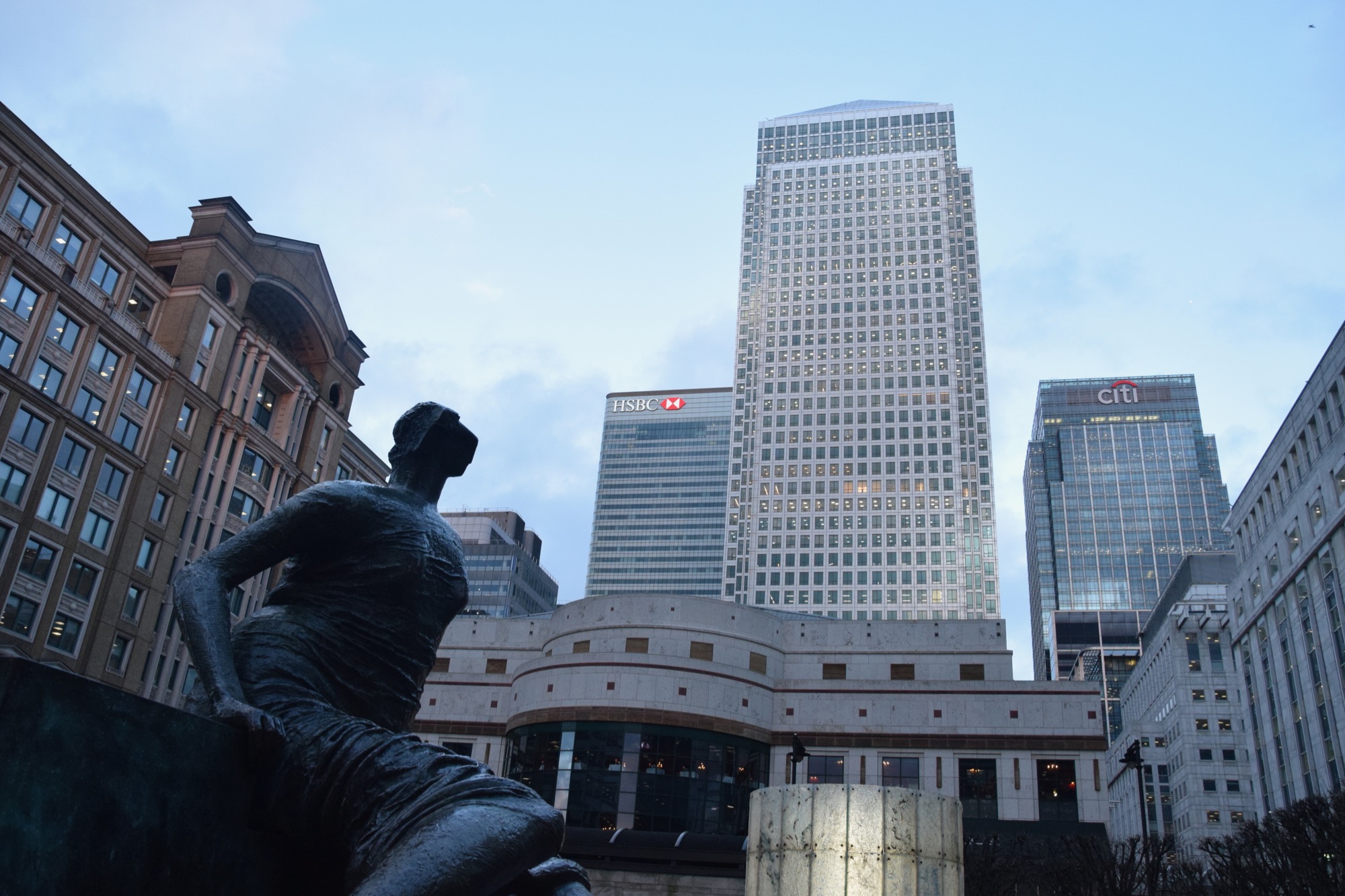 The three central towers of Canary Wharf—One Canda Square, and the HSBC and Citi buildings behind it—dominate the photo. Other office buildings surround, beneath a dim sky. In the foreground is a statue of a seated woman draped in a cloth, her head abstracted.
