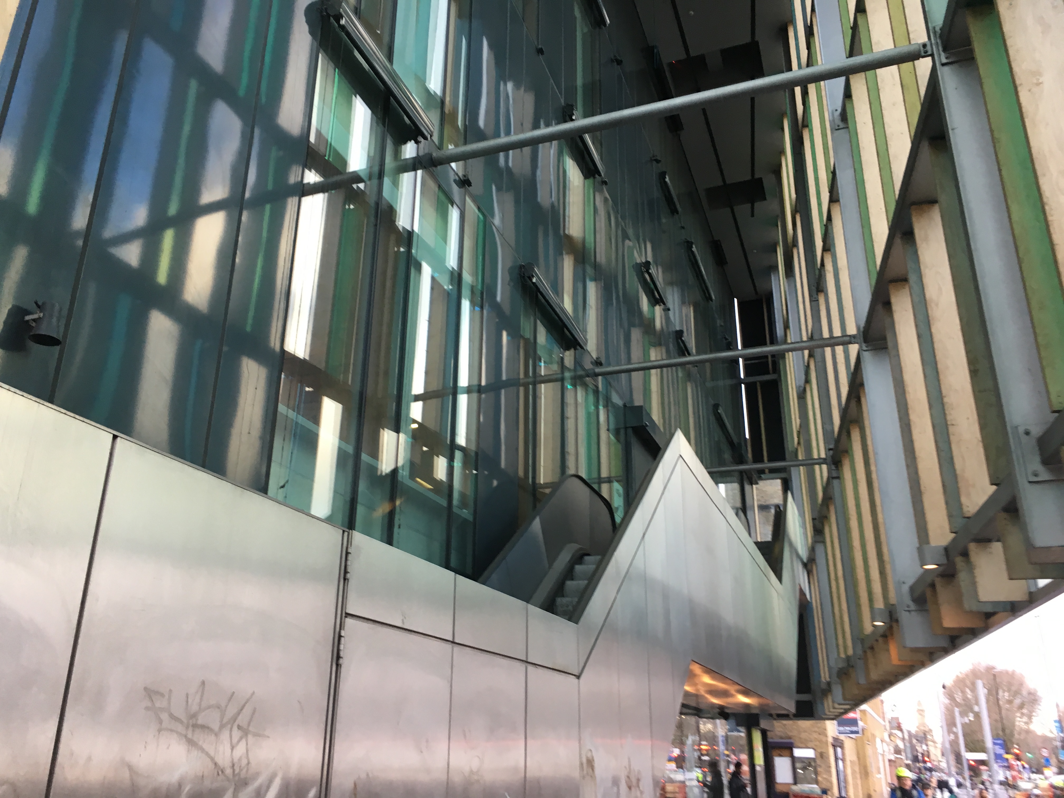 Looking up between a building’s false front and its real outer wall; both are glass, with green and turquoise accents. Leading up in the gap between the two is a series of escalators, the access to which is shut off by metal hoardings. Below the outer wall, a busy street can be seen.
