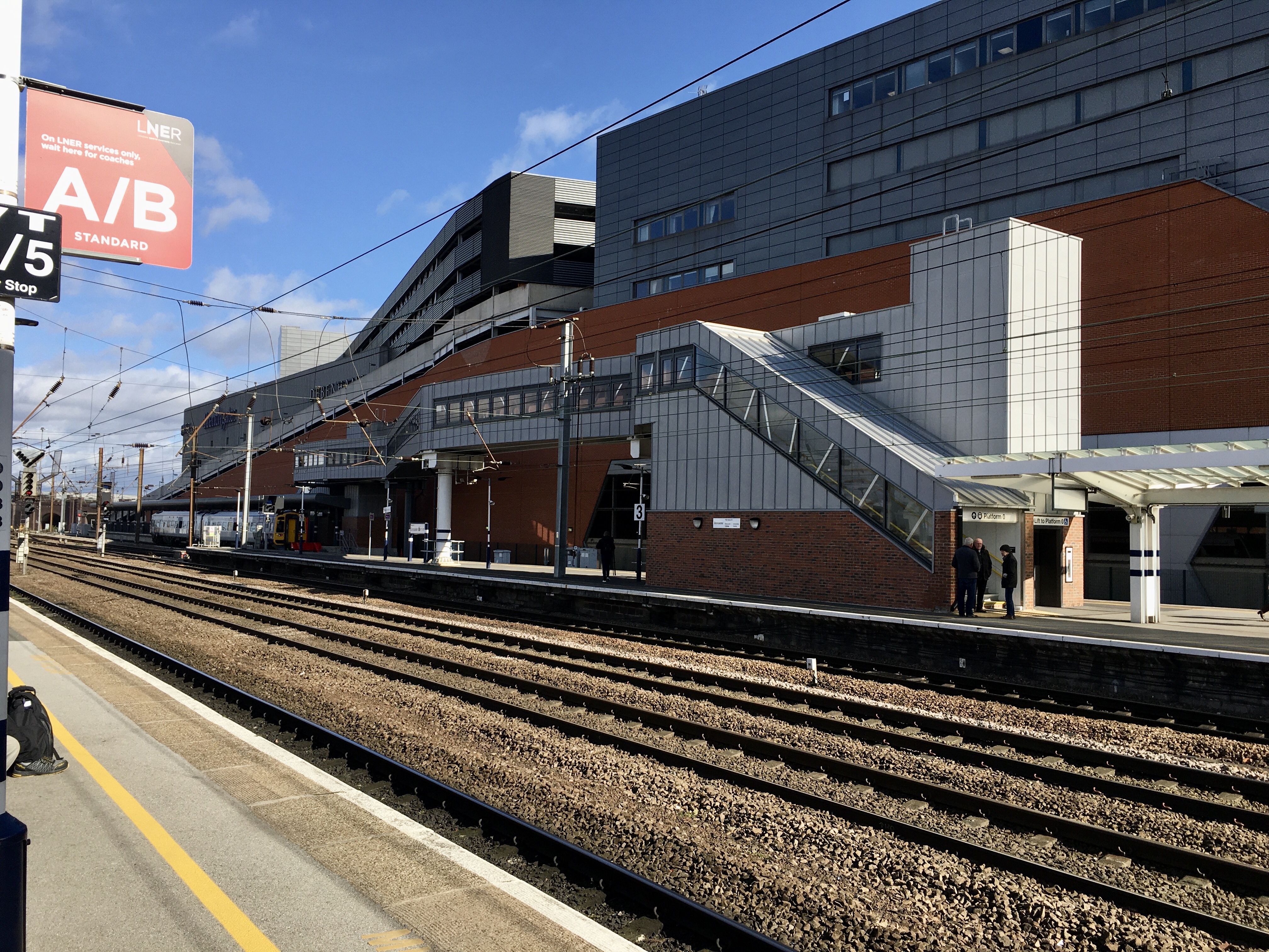 Across a railway line, a platform, on which is a footbridge. The stairs are brick around ground floor height, with silver-ish cladding above. The stairs lead to a long walkway, which leads ultimately to a platform in the distance.