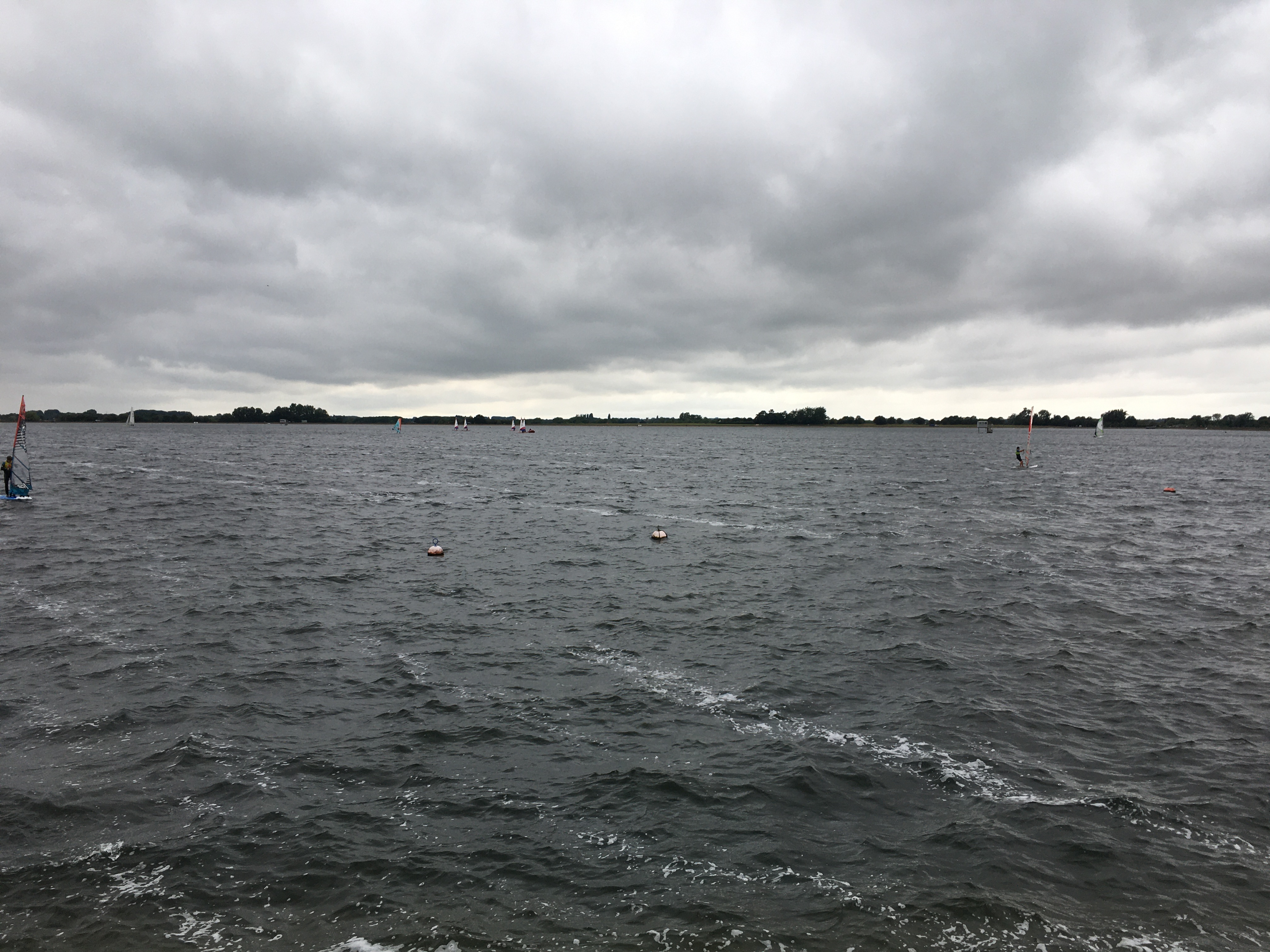 The surface of a large reservoir, with a few people sailing on its surface. The water is grey, as is the sky, except where the latter gets lighter above the horizon. On the horizon itself are lots of tiny silhouettes of trees.
