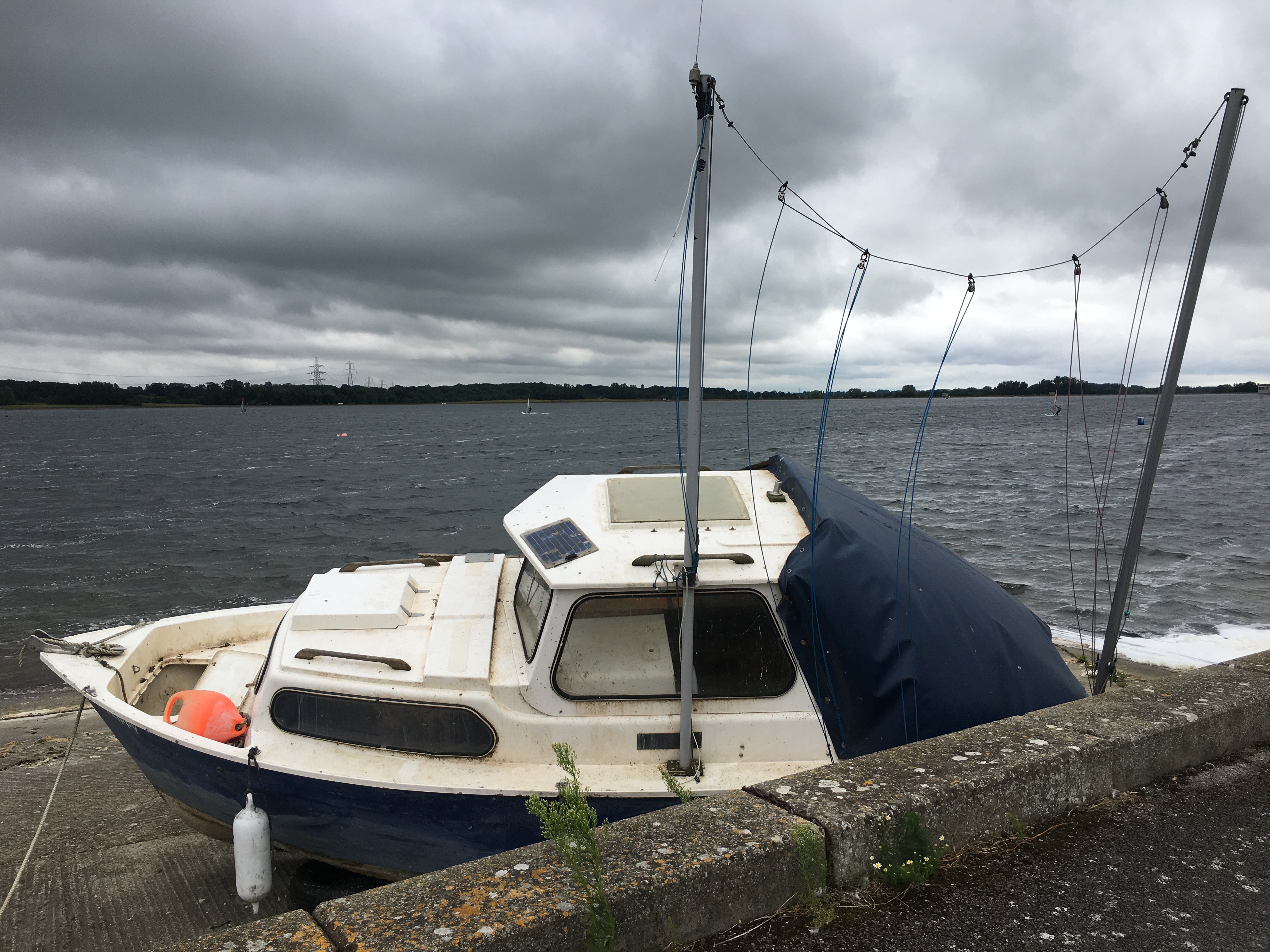 A small motorboat, painted white with a dark blue hull, sitting on a concrete ledge below the path on which the photographer is standing. Behind it is a resvoir with people sailing on it, the water a dark grey matching the overcast sky. In the distance is a wooded area, with pylons sticking out from the top.