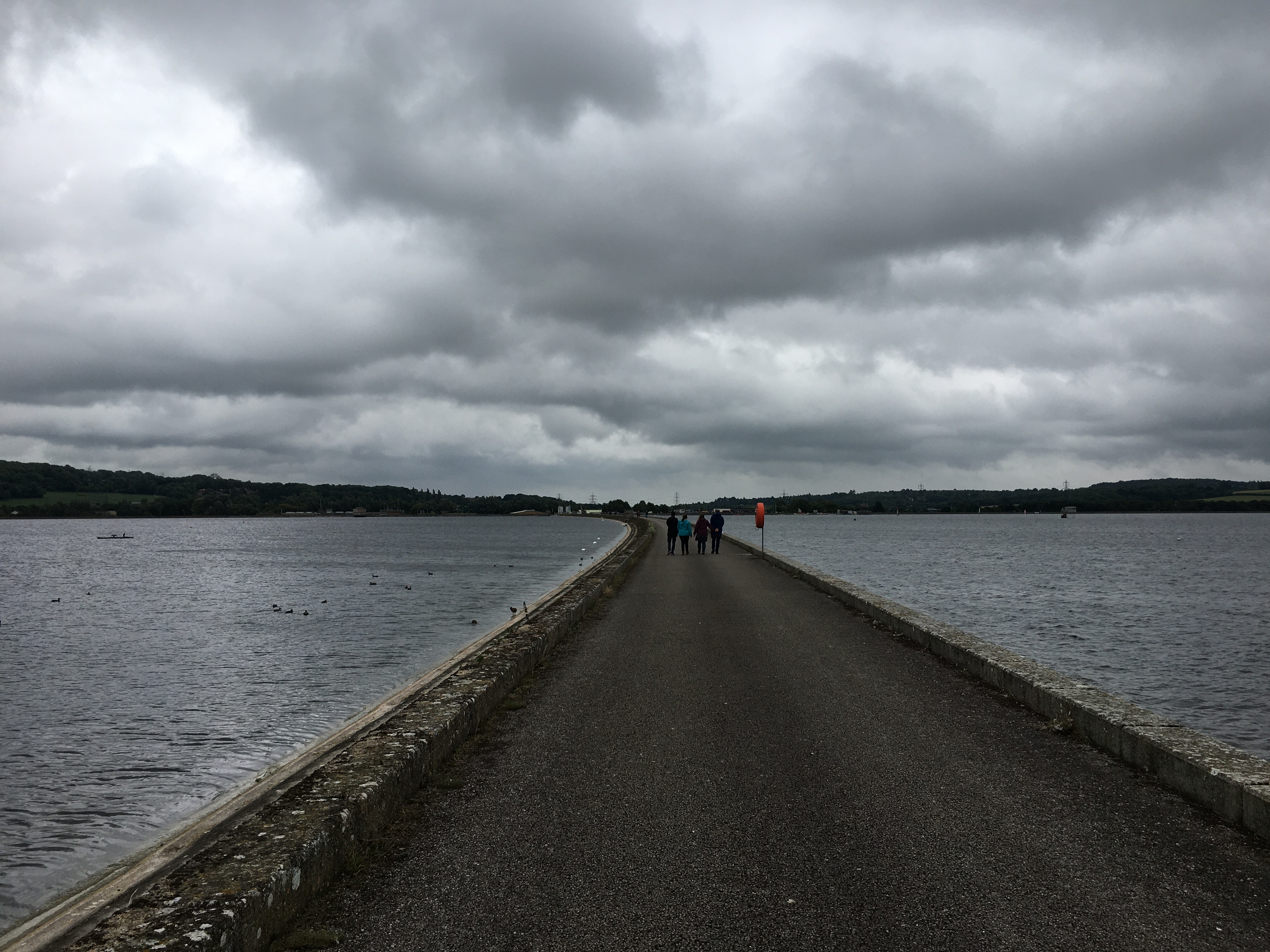 A causeway between two bodies of water. A little further down the causeway from the photographer, next to a life ring, is a group of four people walking side-by-side. In the stance are some hills, forming a gentle V whose apex appears near the end of the causeway. The sky is overcast and dramatically clouded.