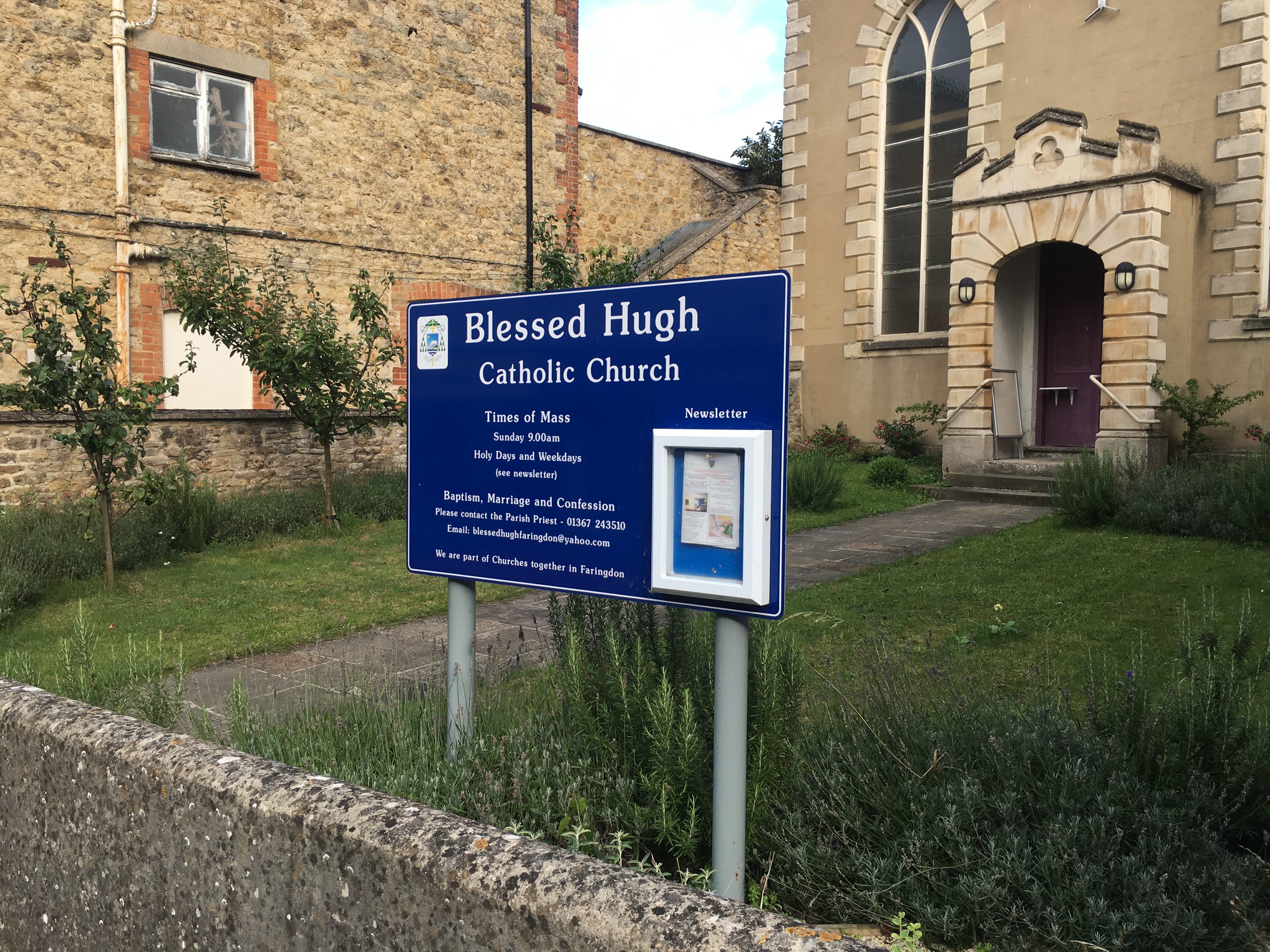 The front garden of a church, with a path between two lawns up to a building with a porch and high arched windows. There's a tall building to the left, and a wall in front of the yard. Behind the wall is a blue sign on metal poles, with a glazed box for the newsletter, the times of mass, and the name of the church: “Blessed Hugh Catholic Church”.