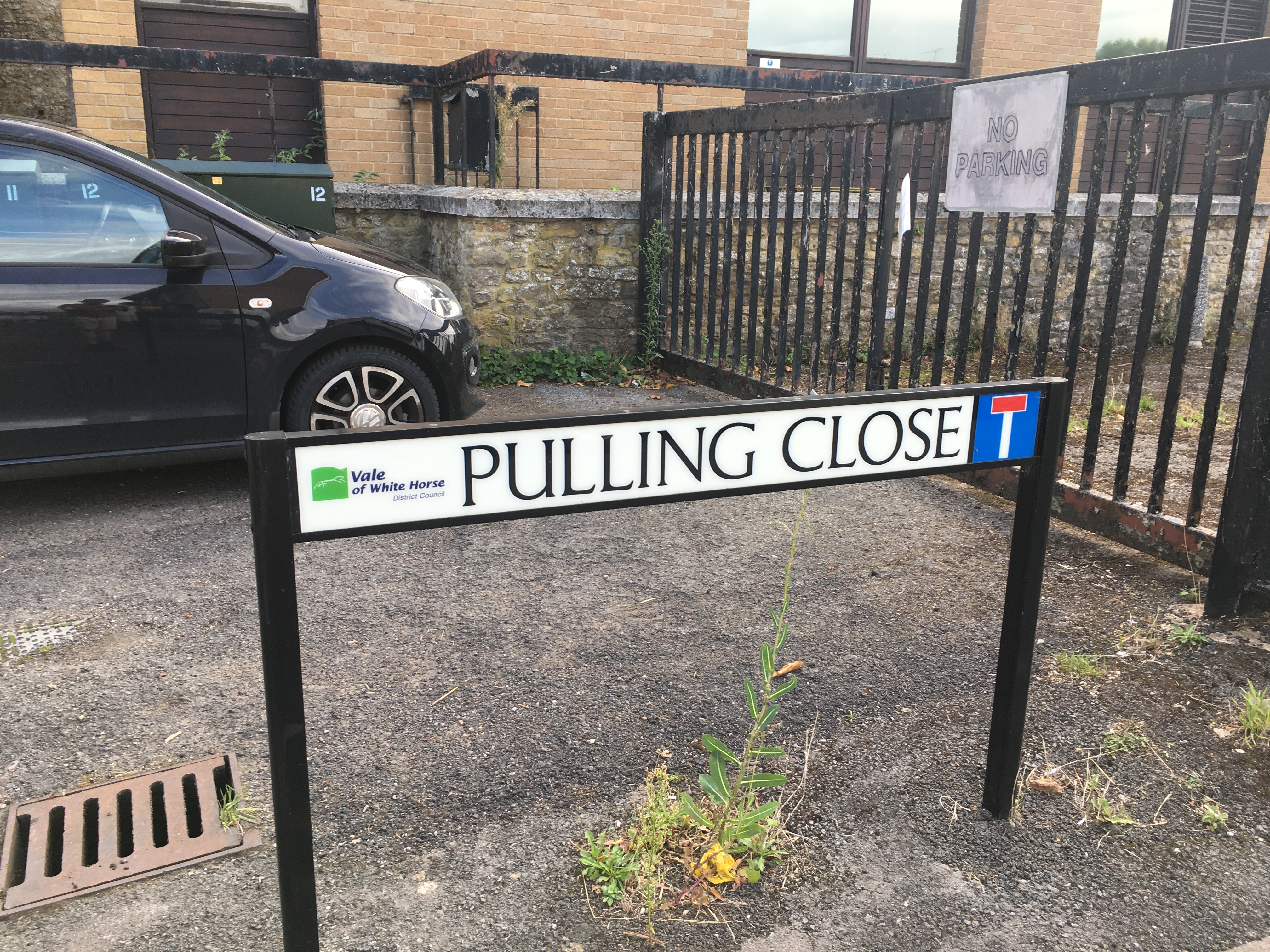 A street sign for “Pulling Close”, with the logo of Vale of White Horse District Council on the left and a cul-de-sac sign on the right. It's in front of a driveway with a parked car on it ahead of some gates with a “No Parking” sign. Behind that is a stone wall that appears to be the foundation of a light-brick modern building with wood-framed windows.