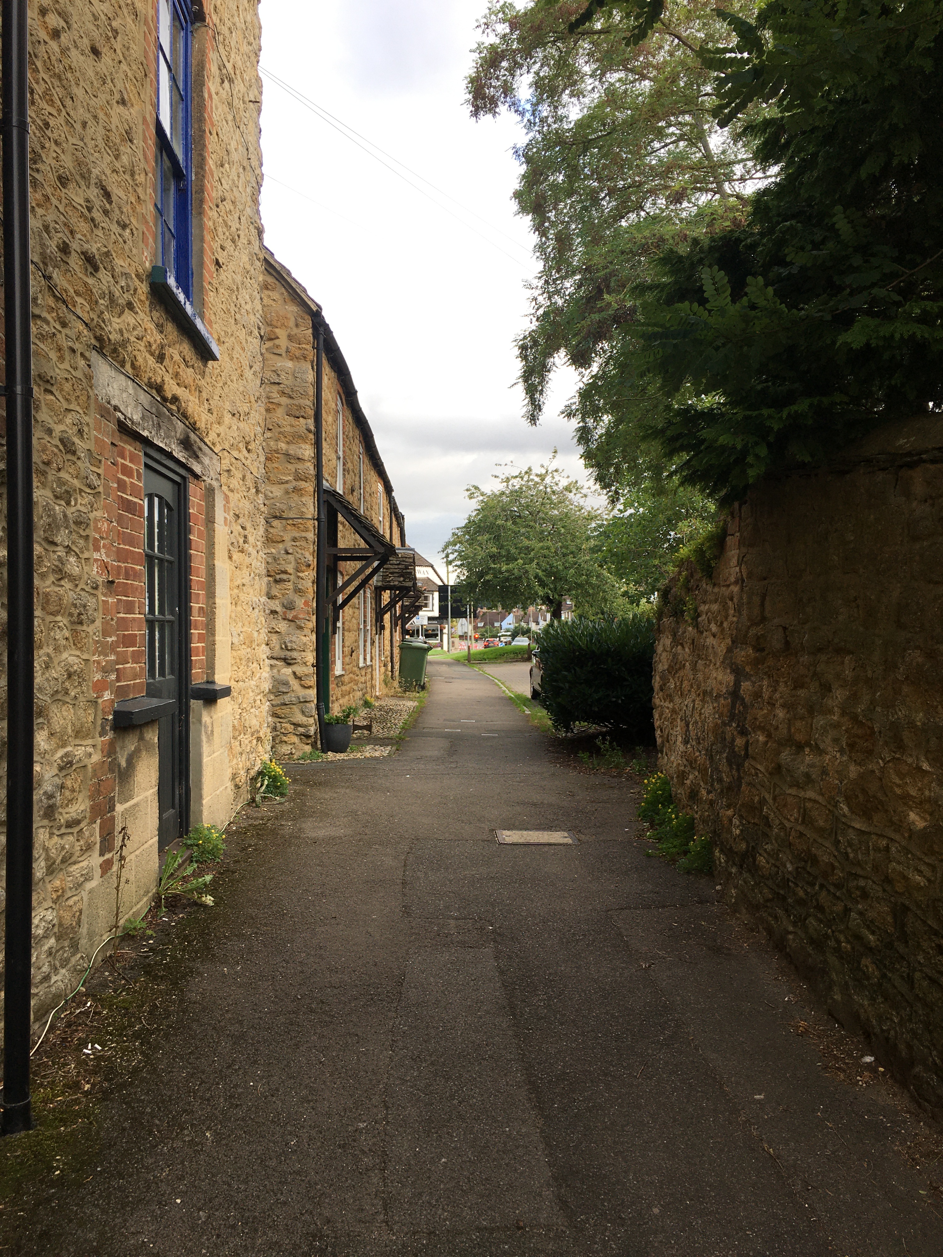 Looking along a path, with stone buildings to the left and a stone wall on the right; the stone wall gives way to a hedge, then a paved area and a tree. The houses to the left have small porches overhanging the doors. There's a road junction in the distance.