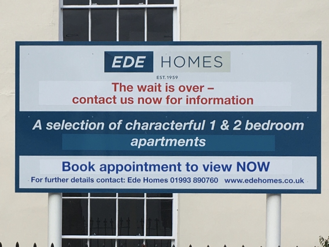 In front of a cream-coloured wall with sash windows divided into a small panes, there is a sign on two white poles. The sign is white, with a dark blue border. It has the logo of a company called “Ede Homes, Est. 1959”, then says in red: “The wait is over – contact us now for information”. Then in white-on-blue: “A selection of characterful 1 & 2 bedroom apartments”. Below that: “Book appointment to view NOW; For further details contact: Ede Homes”, and a telephone number and website are given.