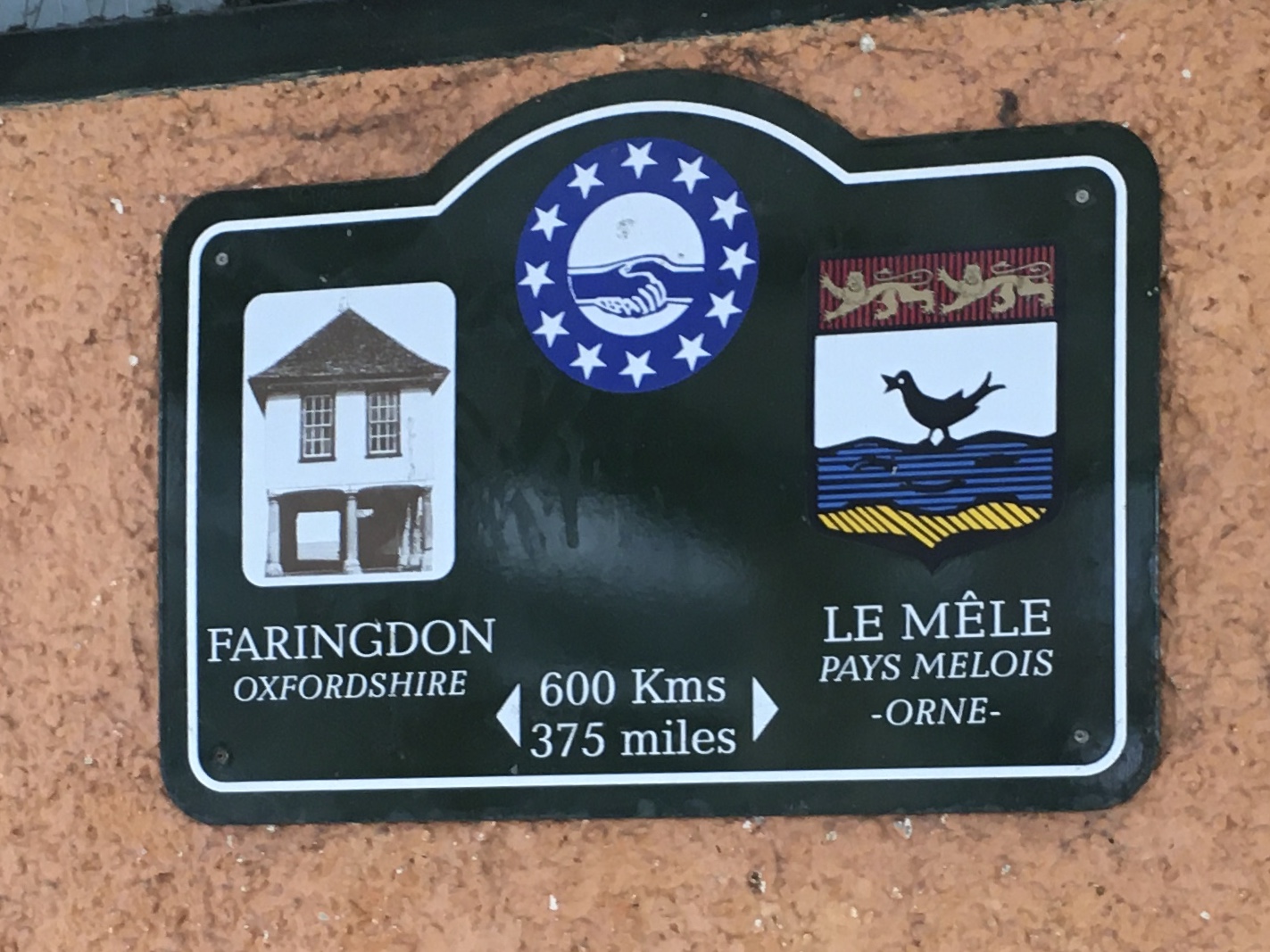 A plaque on an orange-painted pebbledash wall. The plaque is rectangular, with a small hump at the top; it is painted dark green, with a white inset border. The hump is over a depiction of a blue circle containing a ring of twelve white stars, in the centre of which is a white circle containing two blue hands shaking. On either side of the circle, and slightly below it, are representations of two towns. On the left, there is a white rounded rectangle with a brown-coloured representation of an old market building, raised on columns above the street. Below that, in caps, it says “Faringdon, Oxfordshire”. On the right of the blue circle is a shield: the top of this is a red-and-black vertically-striped bar with two lions passant guardant inside, in yellow. The rest of the shield shows a black bird apparently standing on blue-and-black horizonally-striped waves against a white background. Below the waves is a yellow-and-black diagonally-striped representation of sand. Below the shield it says, in white caps, “Le Mêle, Pays Melois, Orne”. At the bottom of the plaque, between the two town representations, is text reading, in white, “600 Kms, 375 miles“; on either side of the text are wedges pointing outwards, to suggest the distance between the towns.