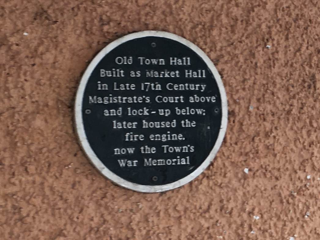 On an orange-painted pebbledash wall, a green circular plaque with a white border. The text of the plaque, in white, says: “Old Town Hall. Built as Market Hall in Late 17th Century. Magistrate‘s Court above and lock-up below; later houses the fire engine, now the Town’s War Memorial”.