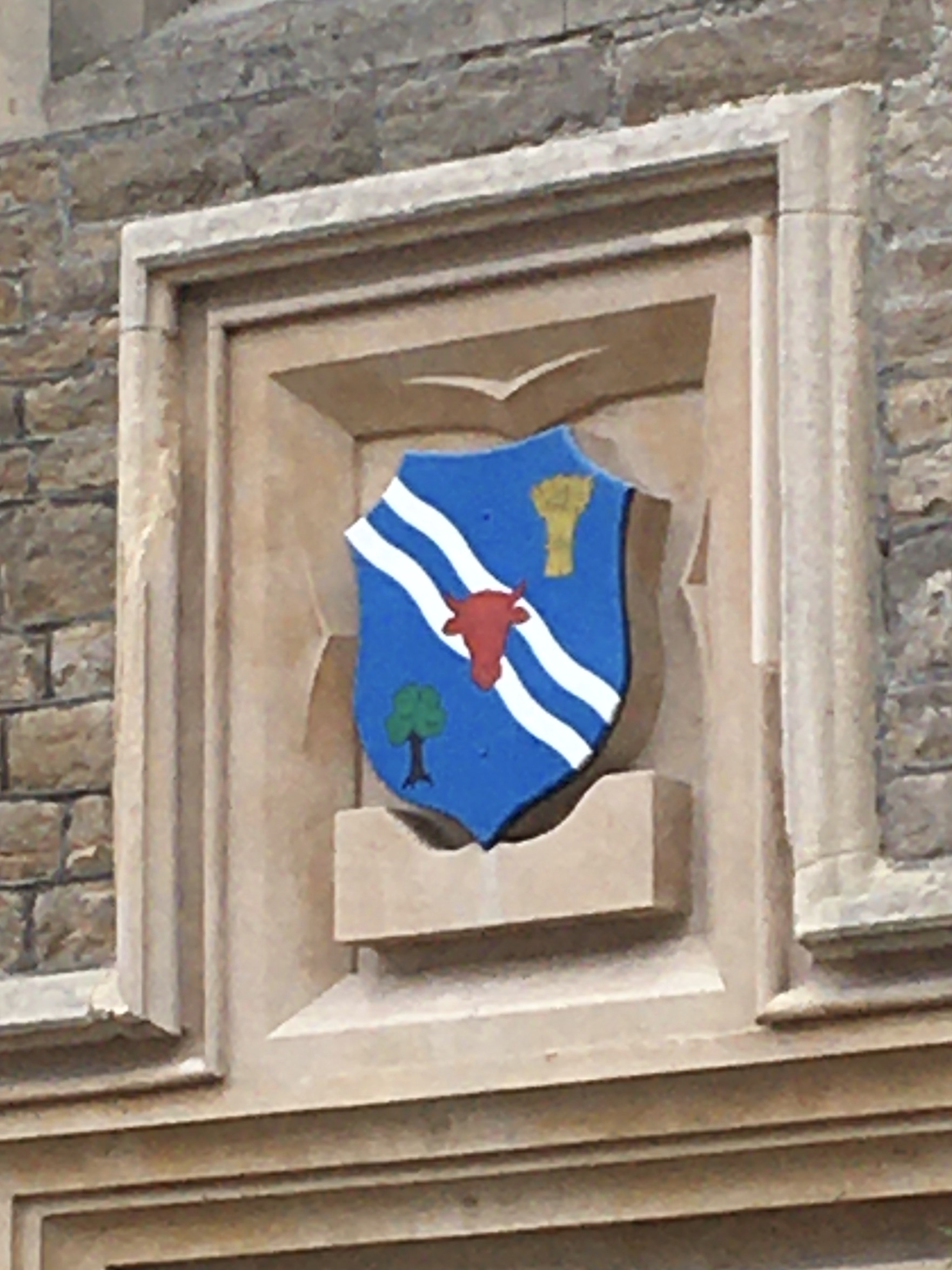 In decorative stonework on a building, the stone forms the shape of a shield. The shield is painted with a heraldic design: blue, with two white diagonal wavy lines from top left to bottom right; atop them, in the middle of the shield, is a red ox’s head facing forwards. At top-right there’s a sheaf of wheat, and at bottom-left a tree.
