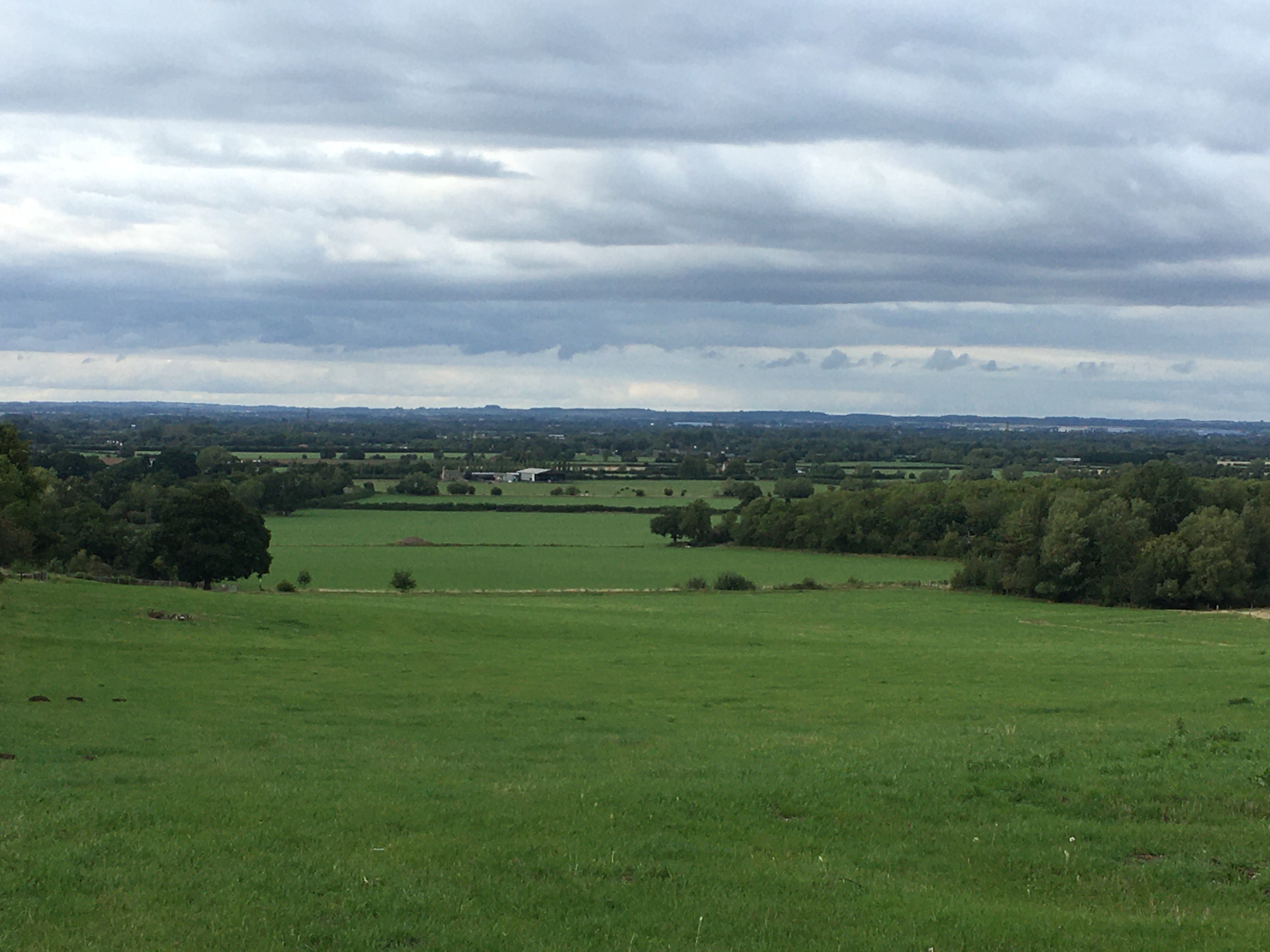 Looking a long distance over fields, with various farm buildings and trees visible, towards low hills in the distance. The sky is fairly overcast, with clouds in left-right bands across the sky.