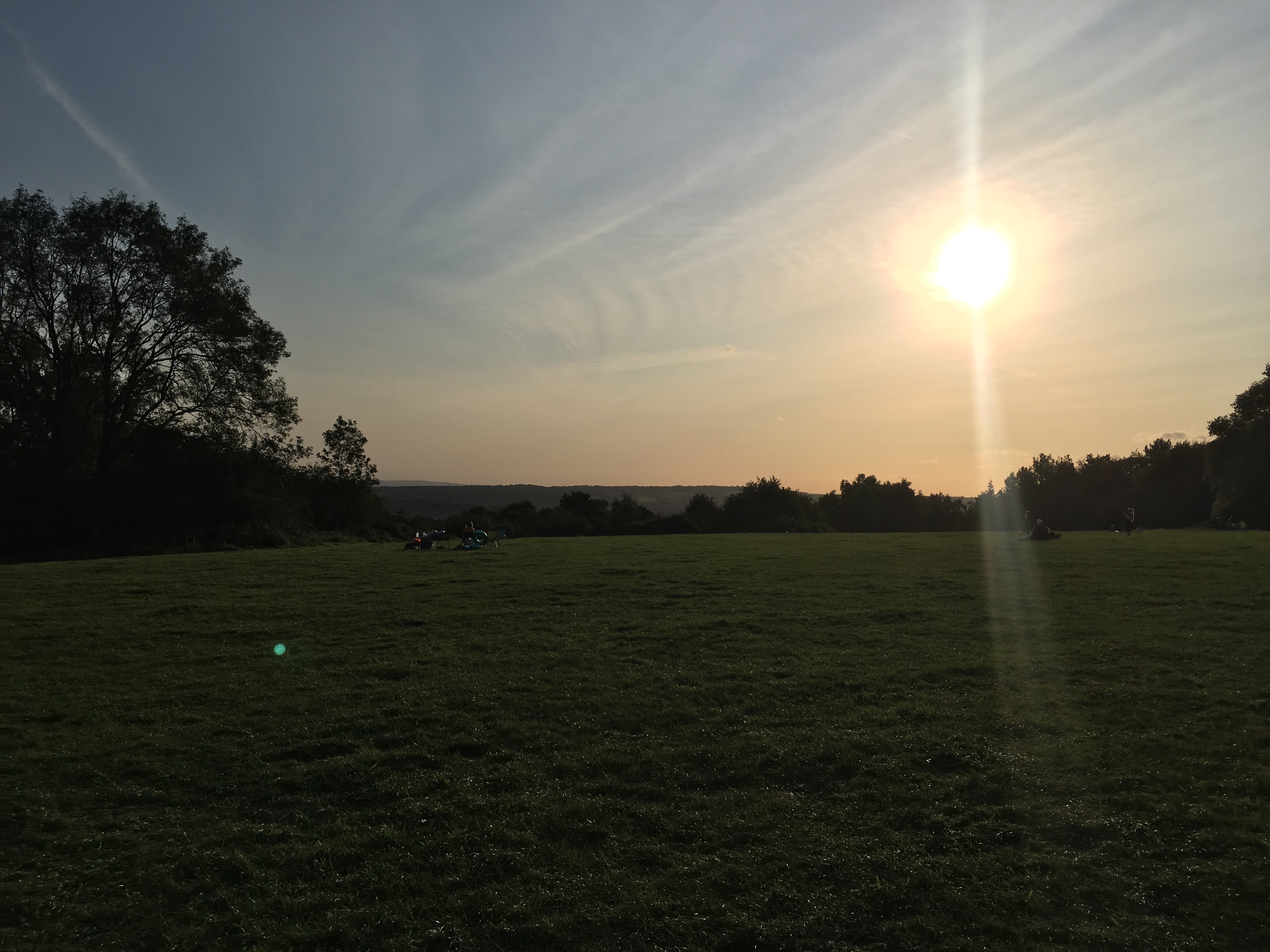 A field at the top of a hill, surrounded by trees silhouetted against the light from the sun, which appears at the upper-right of the picture. Low hills are on the horizon, beneath a blue-yellow sky streaked with clouds.