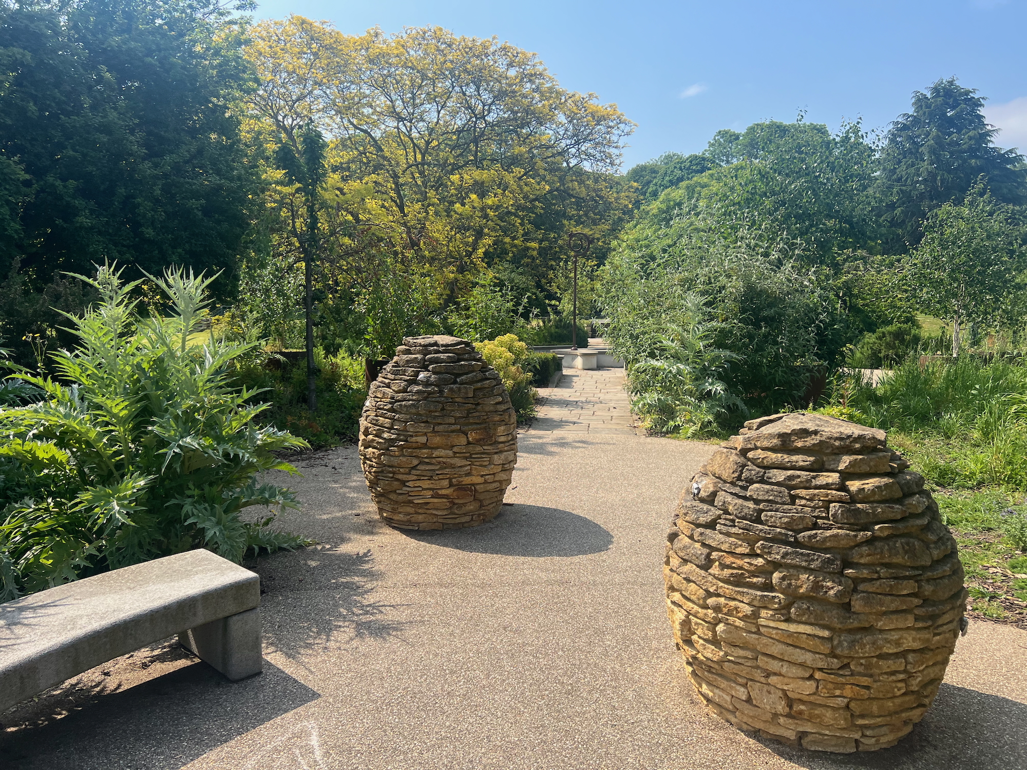 A formal garden in a park. In the foreground are two egg-shaped structures built with stones that look like they've come from the abbey ruins nearby, which are about a metre high. The structures are staggered, the right one in front of the left, with the path undulating as it recedes into the background. A variety of different trees and shrubs surround them. It's a sunny day.