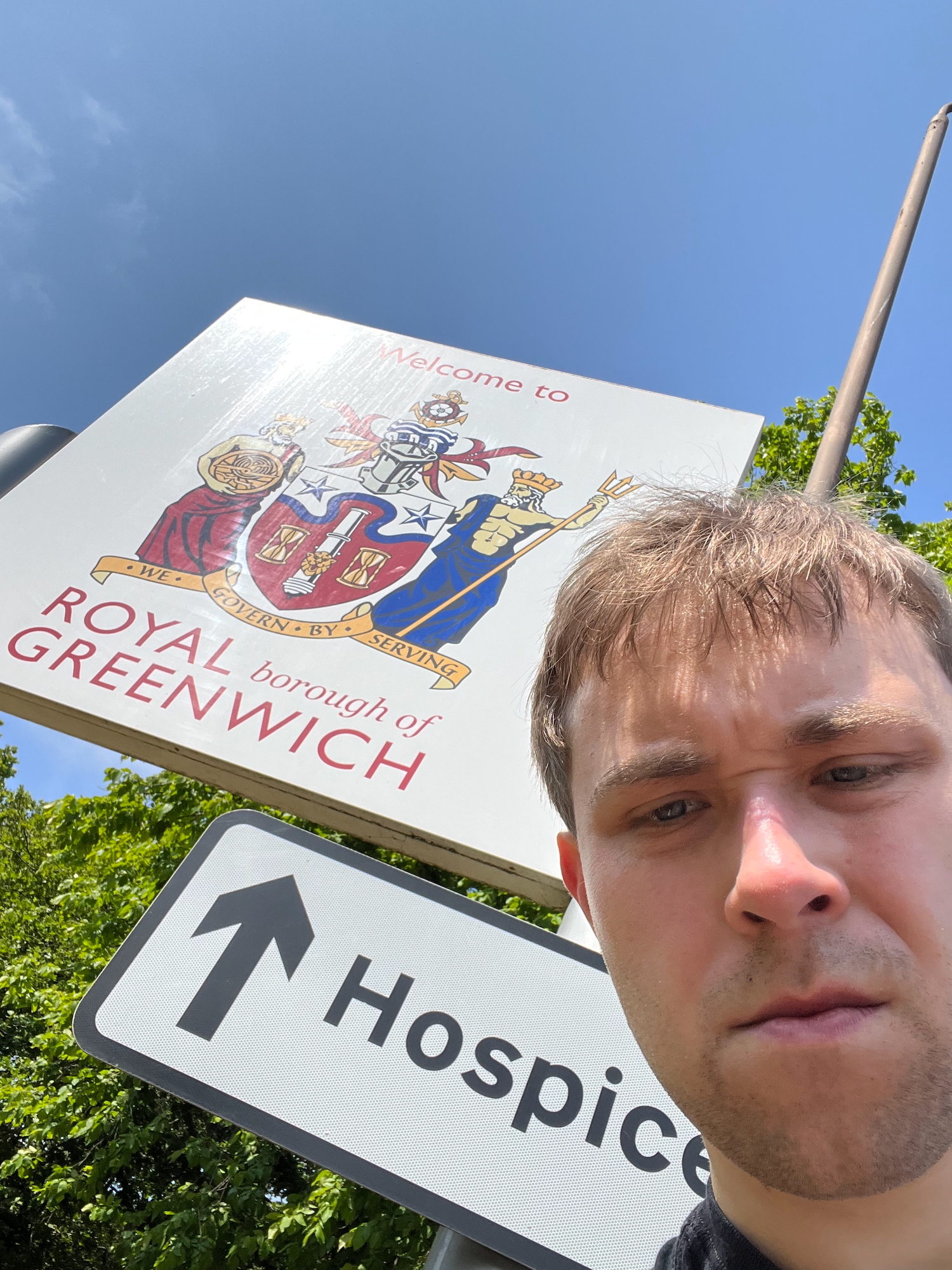 A selfie of the blog's author, standing in front of a sign that says "Welcome to the ROYAL borough of GREENWICH". The words "Welcome to" appear above, and the other words appear below, the borough's coat of arms, with motto "We govern by serving". A road sign underneath points straight ahead to a hospice. The sky is almost cloudless, and there is bright green foliage in the background. The author is scowling at the camera.
