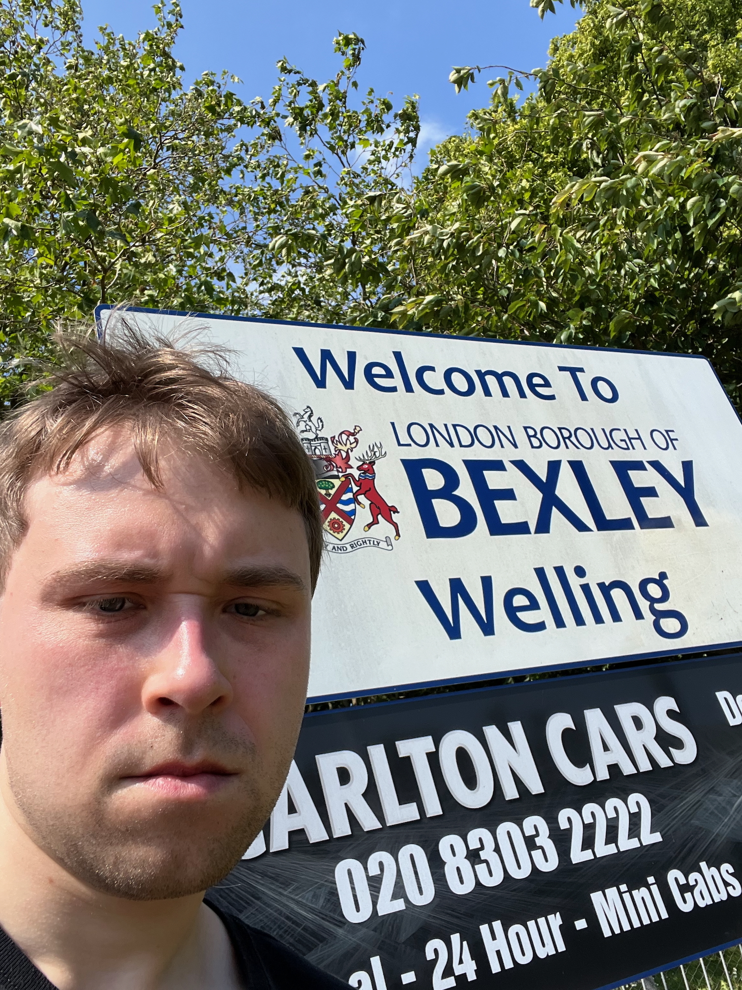 A selfie of the blog's author in front of another sign: "Welcome To LONDON BOROUGH OF BEXLEY, Welling." Underneath the sign is an advert for Carlton Cars, a minicab firm that is open 24 hours. The sky is still almost cloudless, the foliage still bright green, but the author now looks slightly more red-cheeked and disshevelled, even if his scowl is less pronounced.