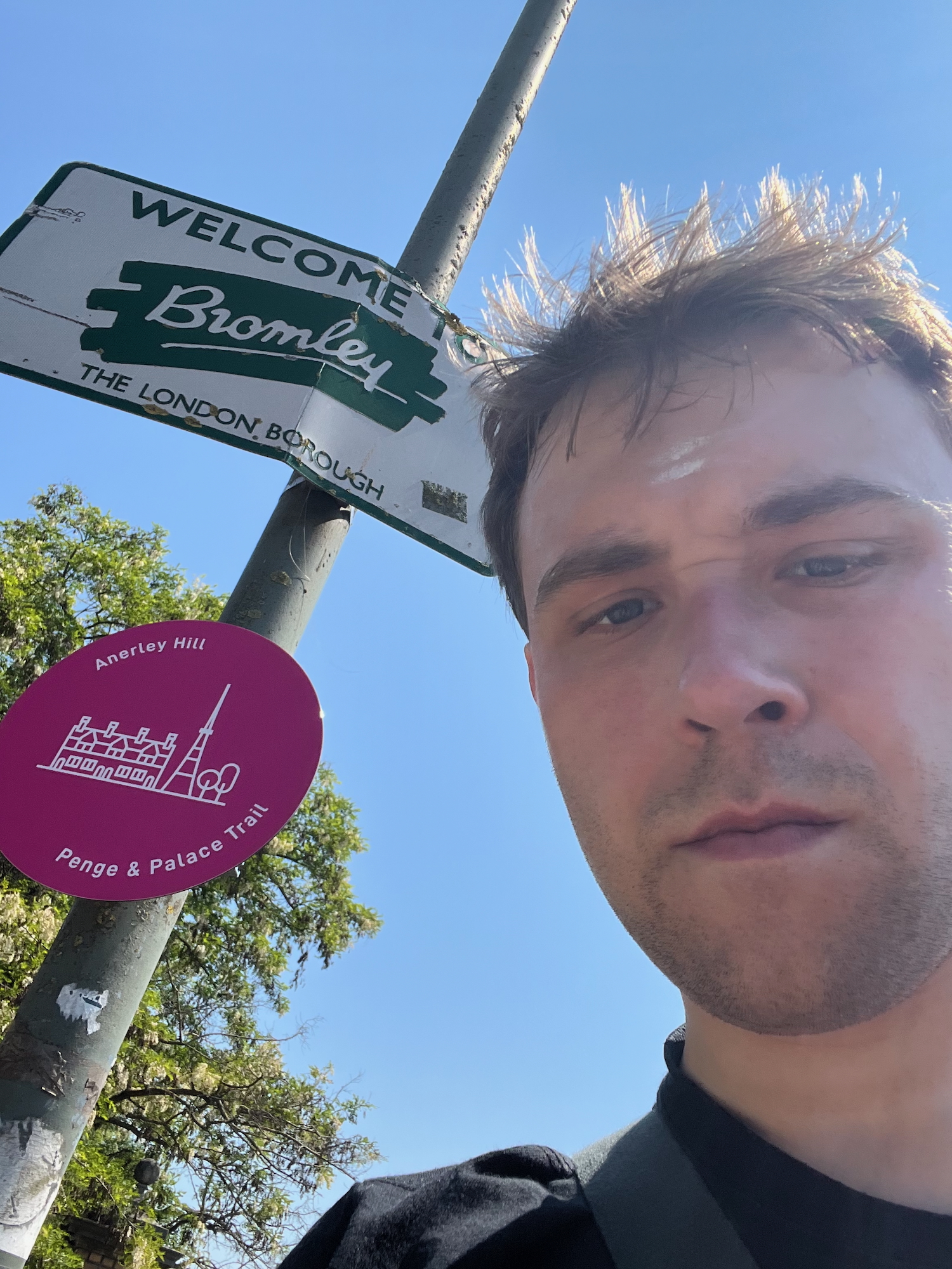 A selfie of the blog's author, peering in from the bottom-right of the photo with sweat shining on his forehead. To the left is a lamppost with two signs, the first reading "WELCOME TO Bromley, THE LONDON BOROUGH"; it is rectangular, with green text on a white background with a green border (except for the name of the borough, which is written in cursive writing in white on a rough green patch). The second sign, smaller and lower down, is circular and pink, with white text around the edge reading "Anerley Hill – Penge & Palace Trail" surrounding a minimalist line drawing of a large house, a transmitter, and two trees. A tree pokes in behind the sign, and the sky, which forms most of the background, is blue from top to bottom.
