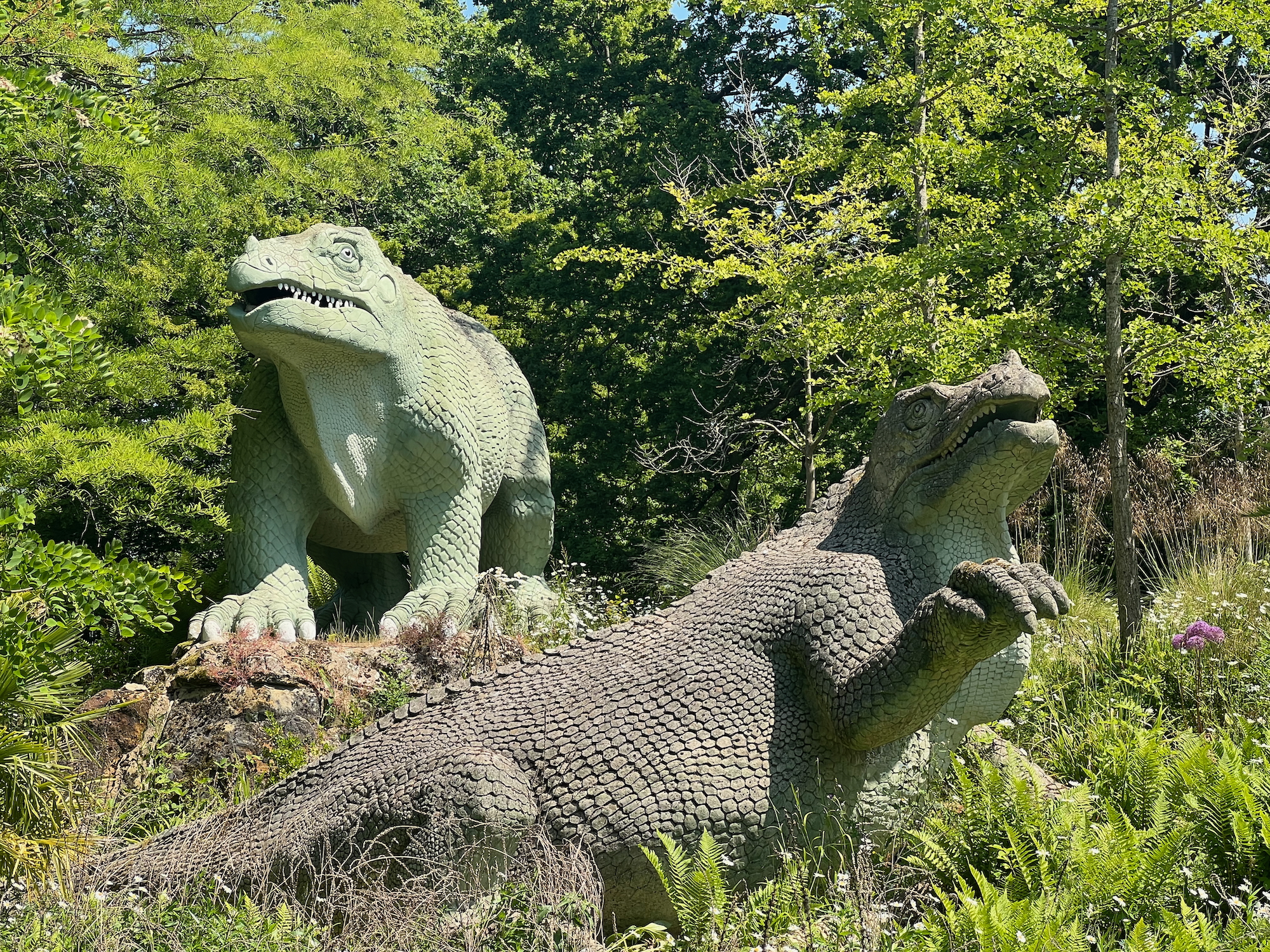 Two sculptures of dinoaurs. The one further back is slightly higher; it is light green, standing on all-fours, and facing the camera. Below it is a brown dinosaur, facing right but with its head turned towards the camera, raising its right foreleg. They sit in the bright sunlight among trees and shrubs typical of a park.