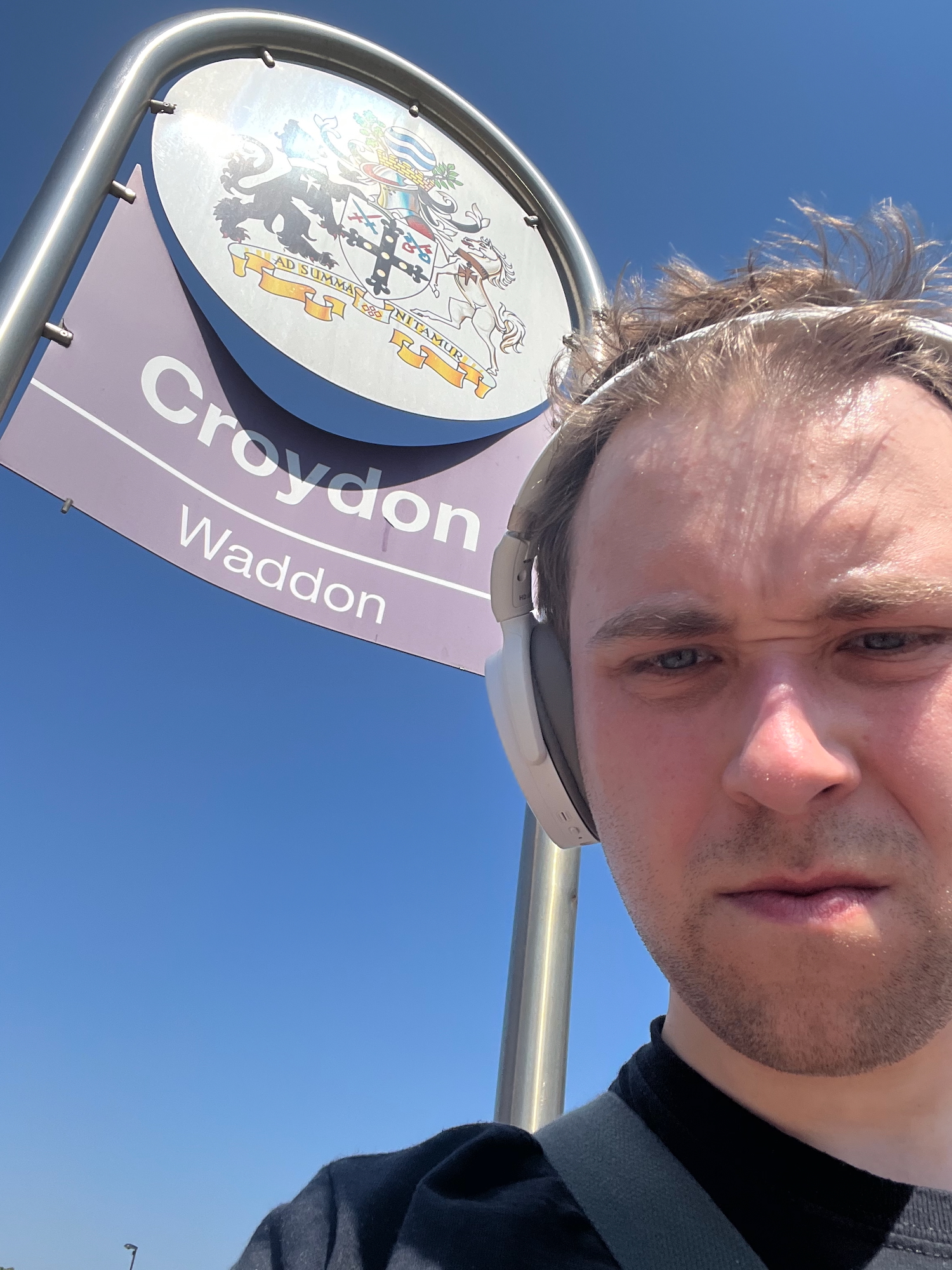 The blog's author, scowling at the camera, his hair messily held off his forehead by a white pair of headphones. He stands in front of a sign made of two parts: a white disc with a coat of arms on it (motto: "AD SUMMA NITAMUR"), and a purple sign which follows the shape of the disk for its concave upper edge, and is a horizontal line at the bottom. The text on the purple part says "Croydon" and (in smaller text) "Waddon", both in white and separated by a white horizontal line. The sign is supported by a single tube of metal, which has been formed into two vertical legs and a semicircular top part to match the disc-shaped sign. The background, other than a distant lamppost just visible at the bottom of the frame, is a blue gradient so featureless it could have been inserted in Photoshop (but wasn't).