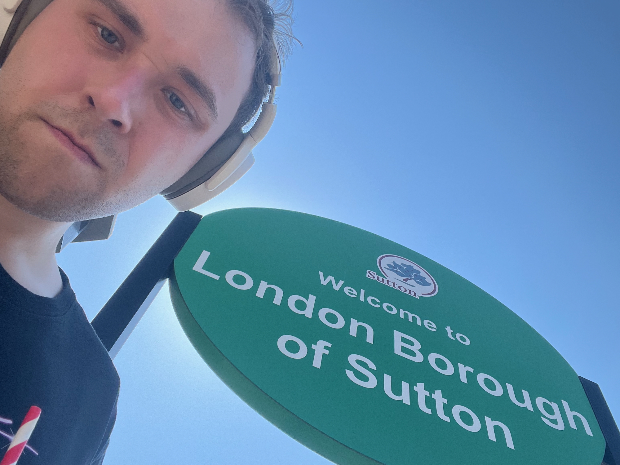 The blog's author looking down at the camera, unsmiling, wearing a black t-shirt and white headphones. Behind him is an elliptical green sign. The main text of the sign is "Welcome to London Borough of Sutton", in white; above this text is the borough's logo, a tree on a white disc with a brown ring near its edge, and the word "Sutton" offset in brown to the lower-left of the tree. The sign is held up by two black posts. Behind the sign is just a blue sky; the sun is masked by the sign, but a halo of its light is visible around the top of the post on the left.